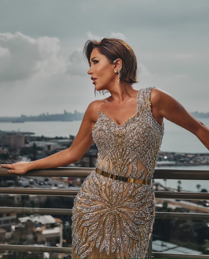 Woman in a sparkling, beaded evening gown standing at a balcony railing with a city and water view behind her (profile pose).