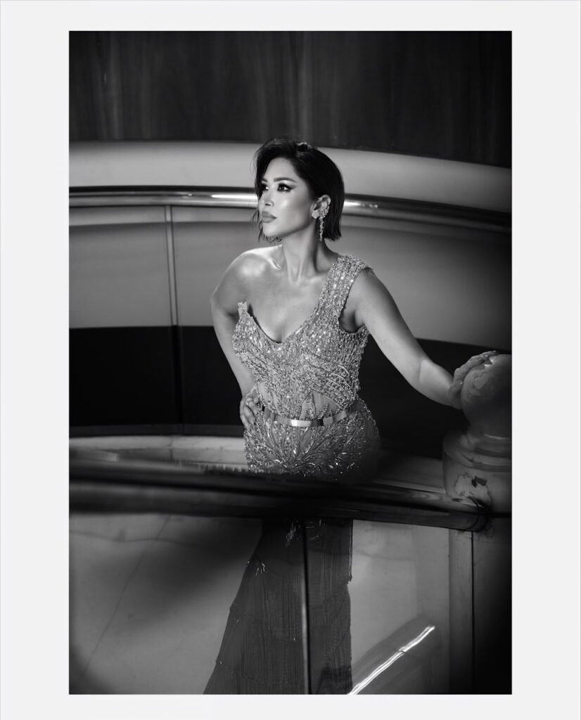 Black-and-white photo of a glamorous woman in a beaded one-shoulder evening gown standing at a railing, looking off to the side.