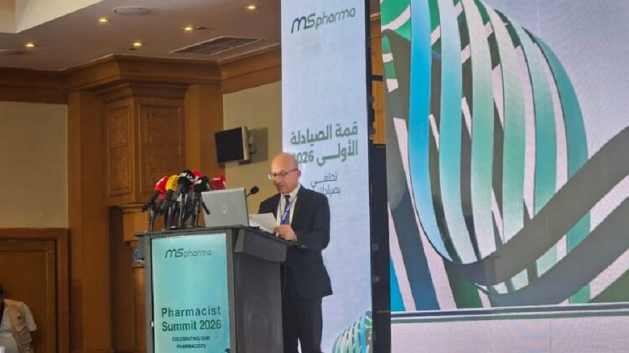 Man in a dark suit speaks at a podium with multiple microphones during a pharmacist summit, with banners and a large screen showing the event branding in the background.