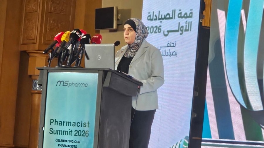 Woman in hijab and gray blazer speaks at podium during Pharmacist Summit 2026, with multiple microphones and a large screen behind her.