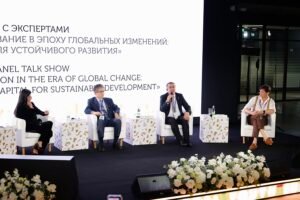 Panel of four speakers seated on a stage with white chairs, a large bilingual screen behind them, and floral arrangements in the foreground.