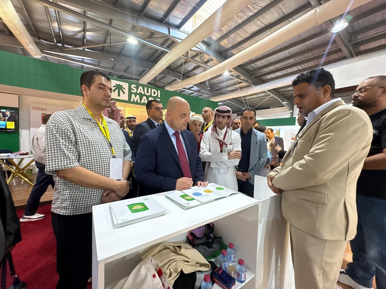 Man in a suit signs documents at a white counter while a group of attendees watches at a trade show booth with green signage behind them, labeled Saudi Map or similar.