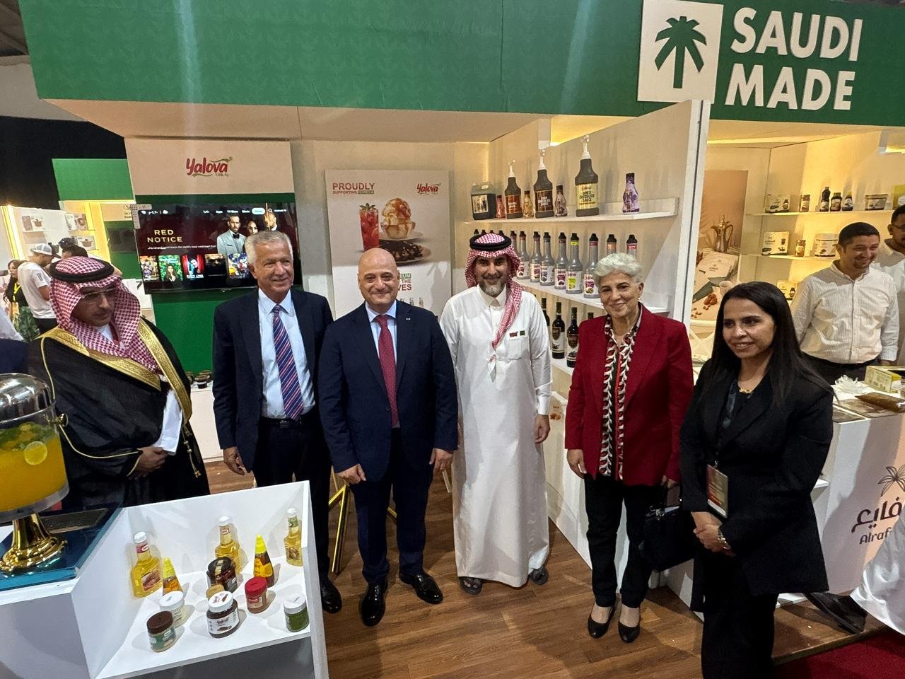 A group of six people standing at a Saudi-made booth with shelves of bottles and jars behind them, posing for a photo at an expo.